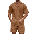 Man wearing a brown patterned shirt and shorts set against a stone wall.